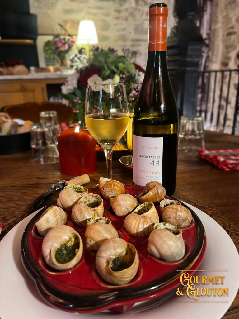 Escargots au beurre persillé servis dans un plat rouge chez Gourmet & Glouton à Chaudes-Aigues, avec un verre de vin blanc en arrière-plan.