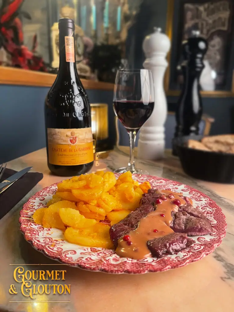 Assiette de biche grillée avec sauce grand veneur, pommes de terre sautées, servie avec un verre et une bouteille de Châteauneuf-du-Pape au restaurant Gourmet & Glouton à Chaudes-Aigues.