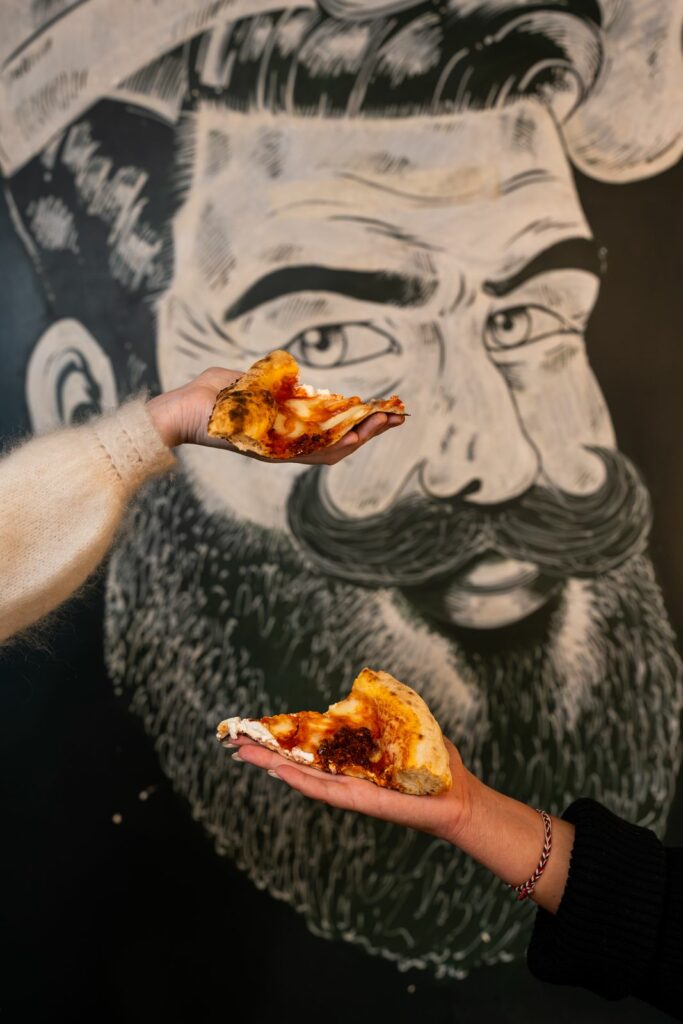 Part de pizza artisanale tenue en main devant une illustration de Pépé Ronchon à Chaudes-Aigues