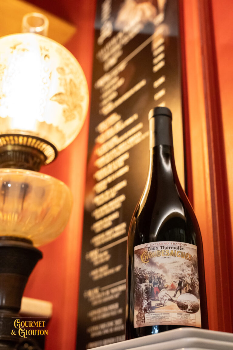 Bouteille « Eaux Thermales de ChaudesAigues » photographiée sur une table du restaurant Gourmet & Glouton, avec une étiquette inspirée d’une affiche ancienne.