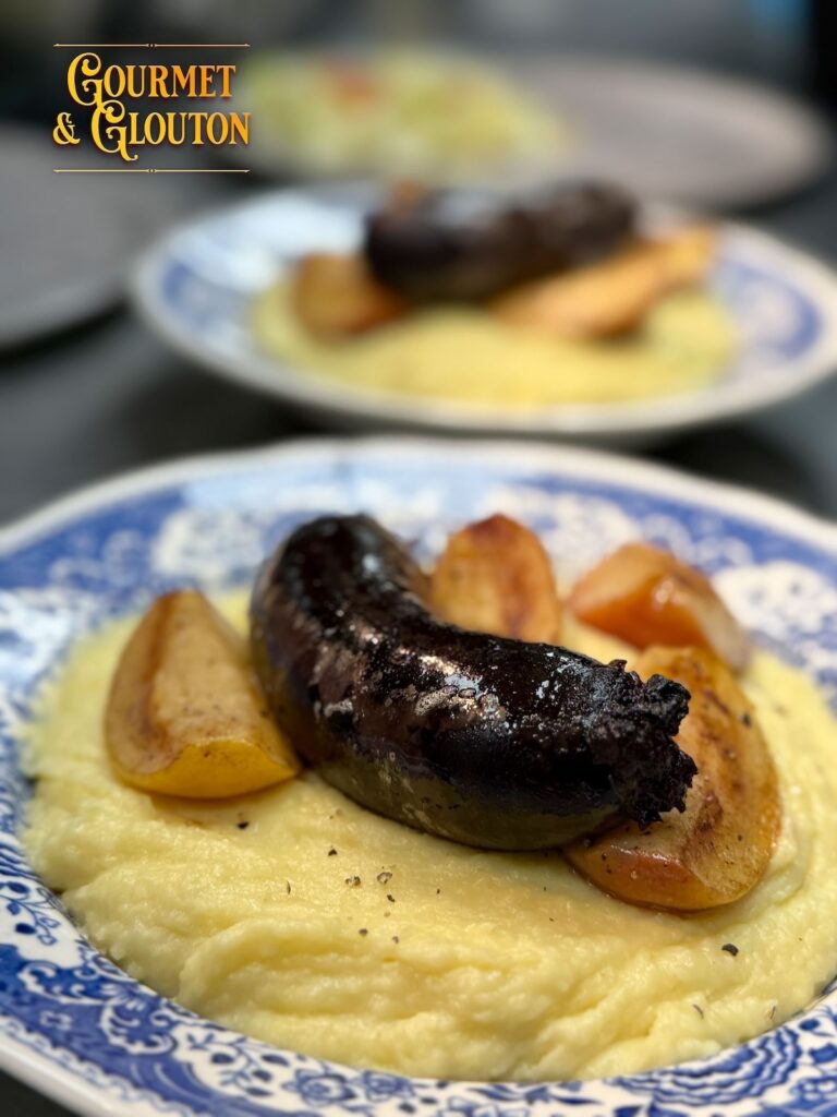 Boudin noir sur une purée maison, entouré de quartiers de pommes dorées, dans une assiette bleue et blanche.