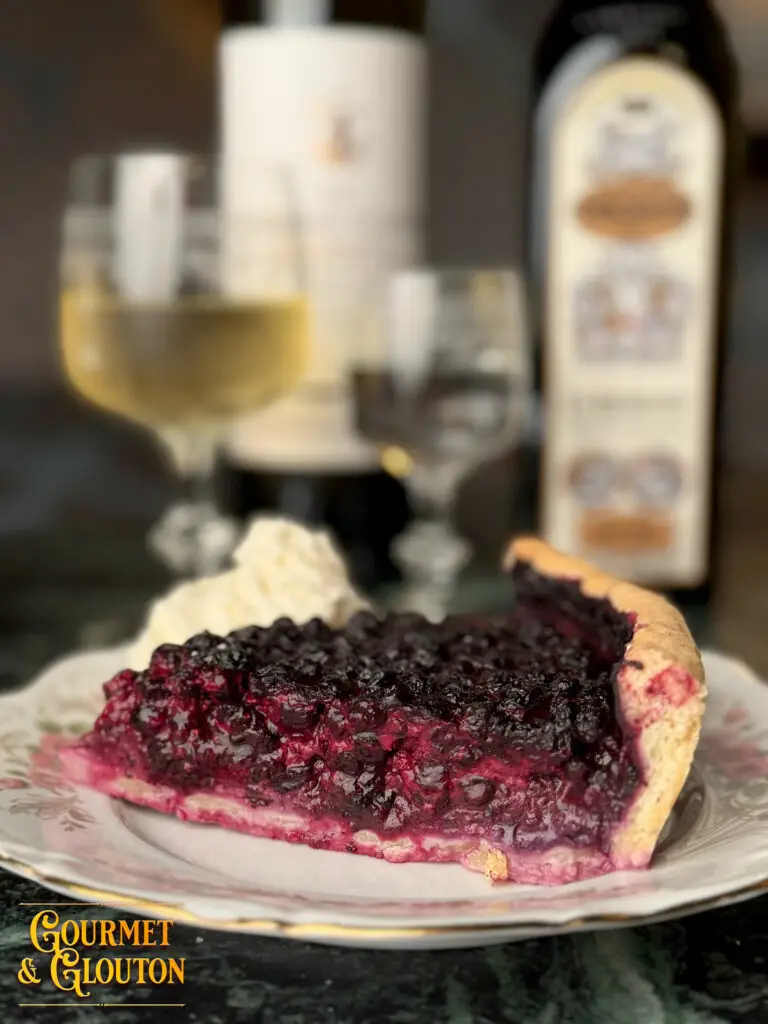 Part de tarte aux myrtilles maison servie avec chantilly, photographiée au restaurant Gourmet & Glouton à Chaudes-Aigues, avec un verre de vin blanc en arrière-plan.