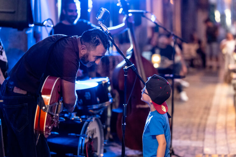 Musicien se penchant vers un enfant devant un micro vintage lors d’un concert en terrasse chez Gourmet & Glouton à Chaudes-Aigues, illustrant un moment de partage et de transmission.