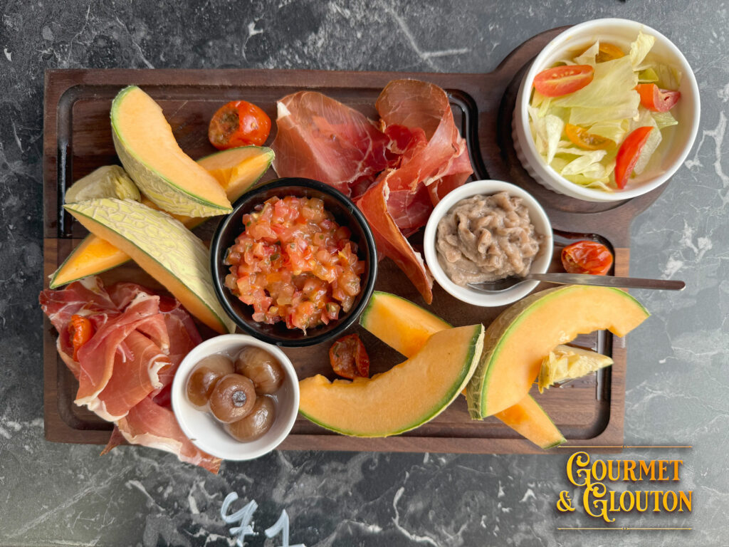 Planche estivale servie chez Gourmet & Glouton à Chaudes-Aigues avec melon, jambon cru, tartare de tomates, caviar d'aubergines, artichaut, oignons Borettane et salade fraîche.