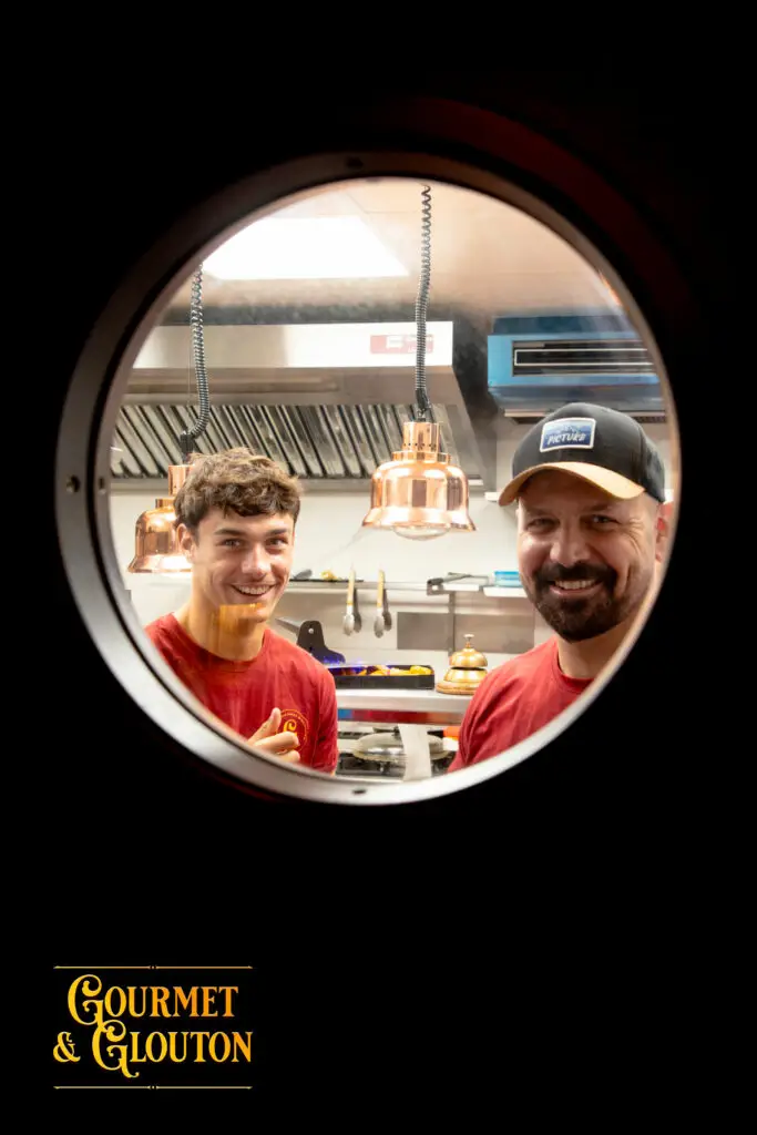 Maxime et Loïc, alias Simon, sourient derrière le hublot de la porte de la cuisine de Gourmet & Glouton.